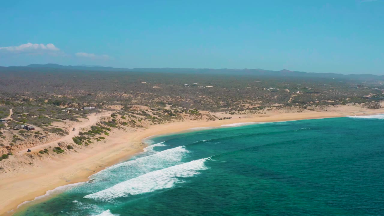 Aerial view over Shipwrecks Beach in Baja California Sur, with turquoise water and white surf meeting a broad sandy shoreline, no visible people.