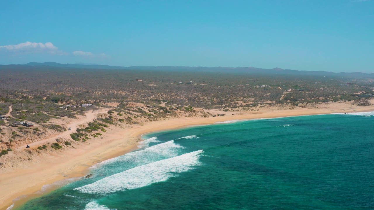 Drone-like aerial frame over Shipwrecks Beach showing surf washing onto an empty sandy shore, with low dunes, scattered greenery, and inland tracks in the background, no visible people.