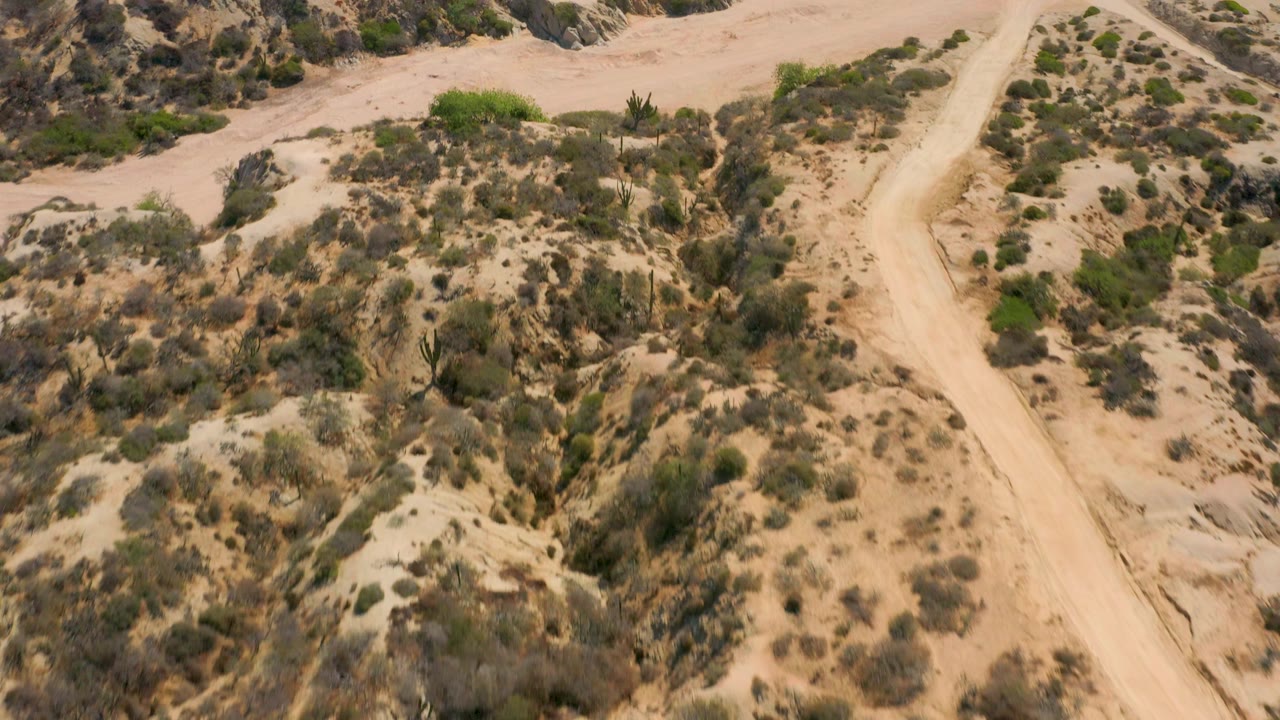 Overhead view at Shipwrecks Beach, Baja California Sur, with a sandy wash, low scrub-covered slopes, and a dirt path near the right edge, with no visible people.