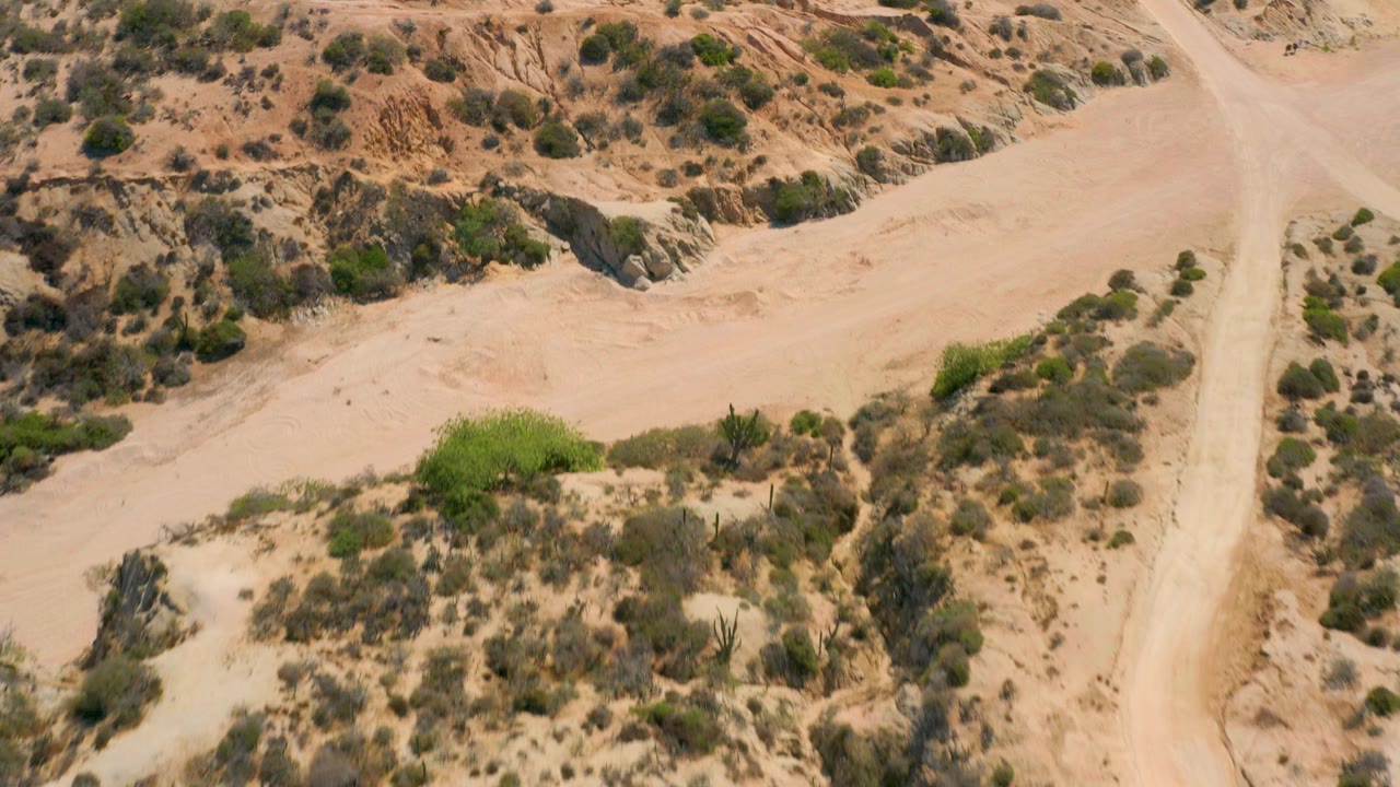 Aerial frame over Shipwrecks Beach, Baja California Sur, showing a pale sandy wash, a vertical dirt track, eroded tan ground, scattered shrubs, and no visible people.