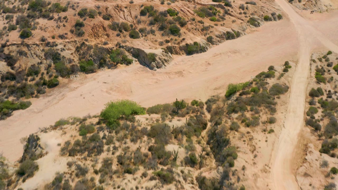 Aerial view at Shipwrecks Beach with a broad pale sand channel crossing eroded desert terrain, green shrubs clustered near the wash, and no visible people.