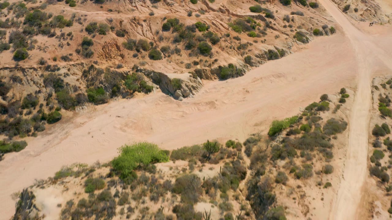 High aerial frame over Shipwrecks Beach, Baja California Sur, with a dirt road running near the right side, a sandy wash across the center, scrub patches, and no visible people.