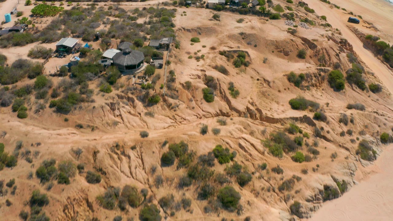 Aerial frame over Shipwrecks Beach in Baja California Sur showing eroded tan bluffs, scattered scrub, small beachside buildings, and no visible people.