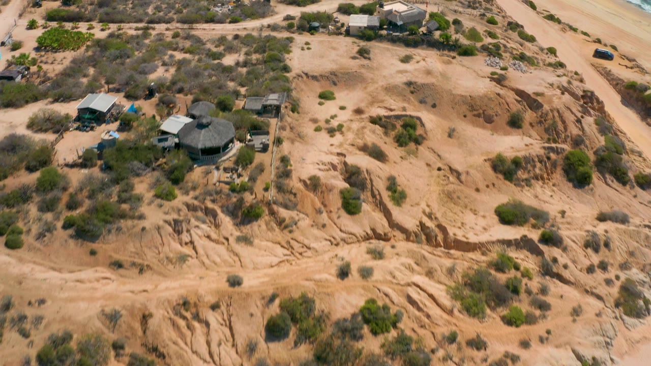 Shipwrecks Beach aerial frame with small coastal buildings, a pale sand road, dry scrubland, a strip of surf at the right edge, and no visible people.