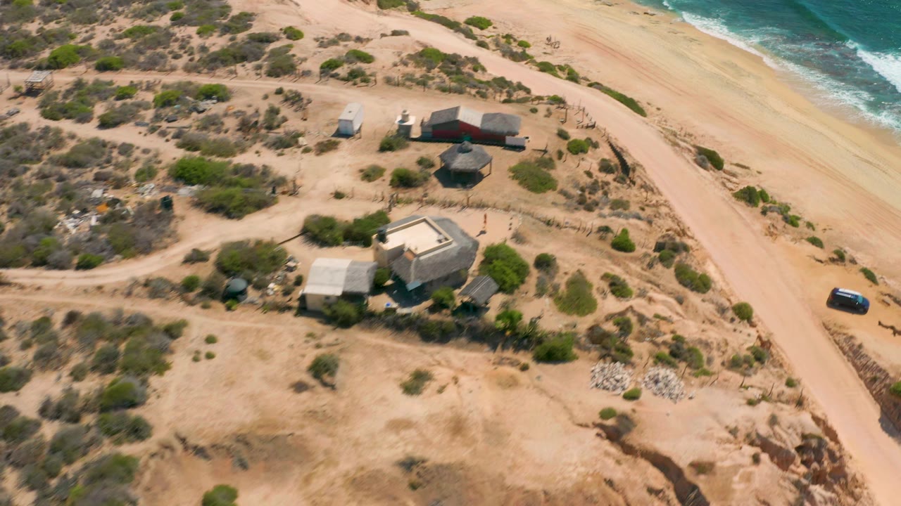 Aerial frame at Shipwrecks Beach with the houses centered lower in view, a dirt road running beside the beach, blue-green water at the top right, and no visible people.
