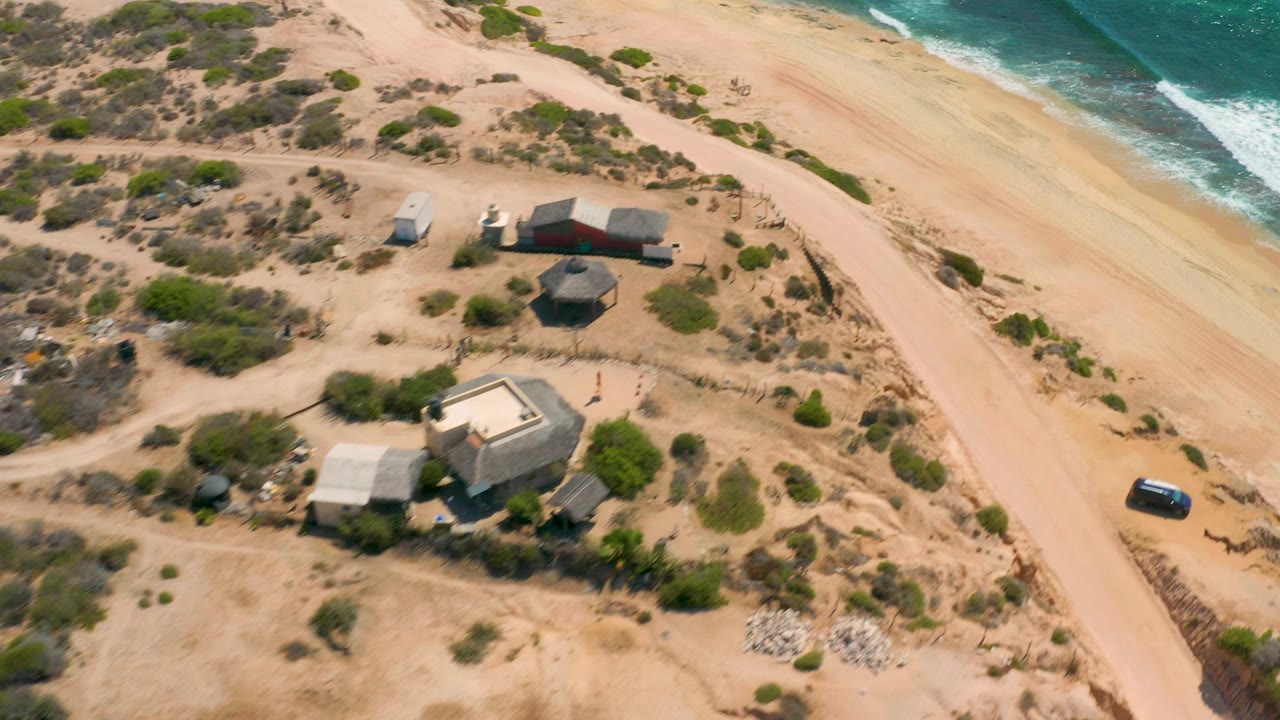 Oblique drone view of Shipwrecks Beach, Baja California Sur, with clustered small buildings, low green shrubs, broad sand, surf breaking along the upper right edge, and no visible people.