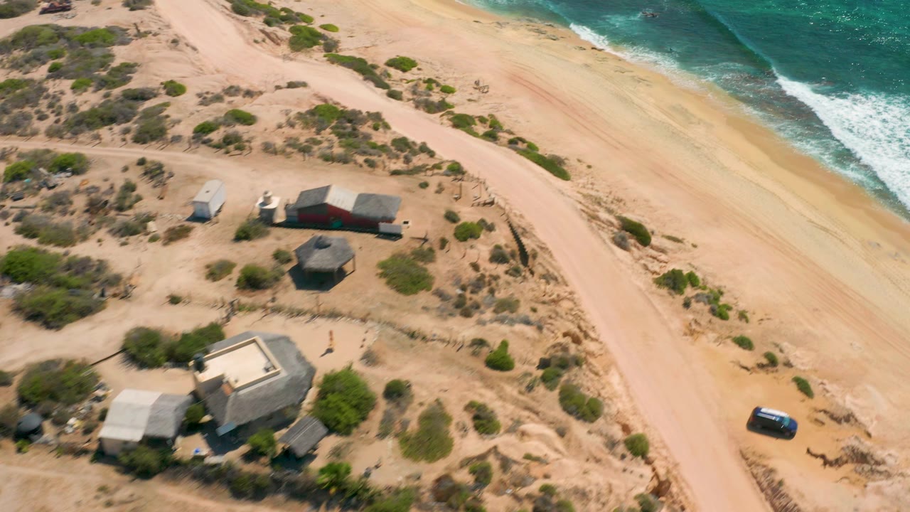 Drone angle over beach houses, dirt road, shrubs, sand, and surf.