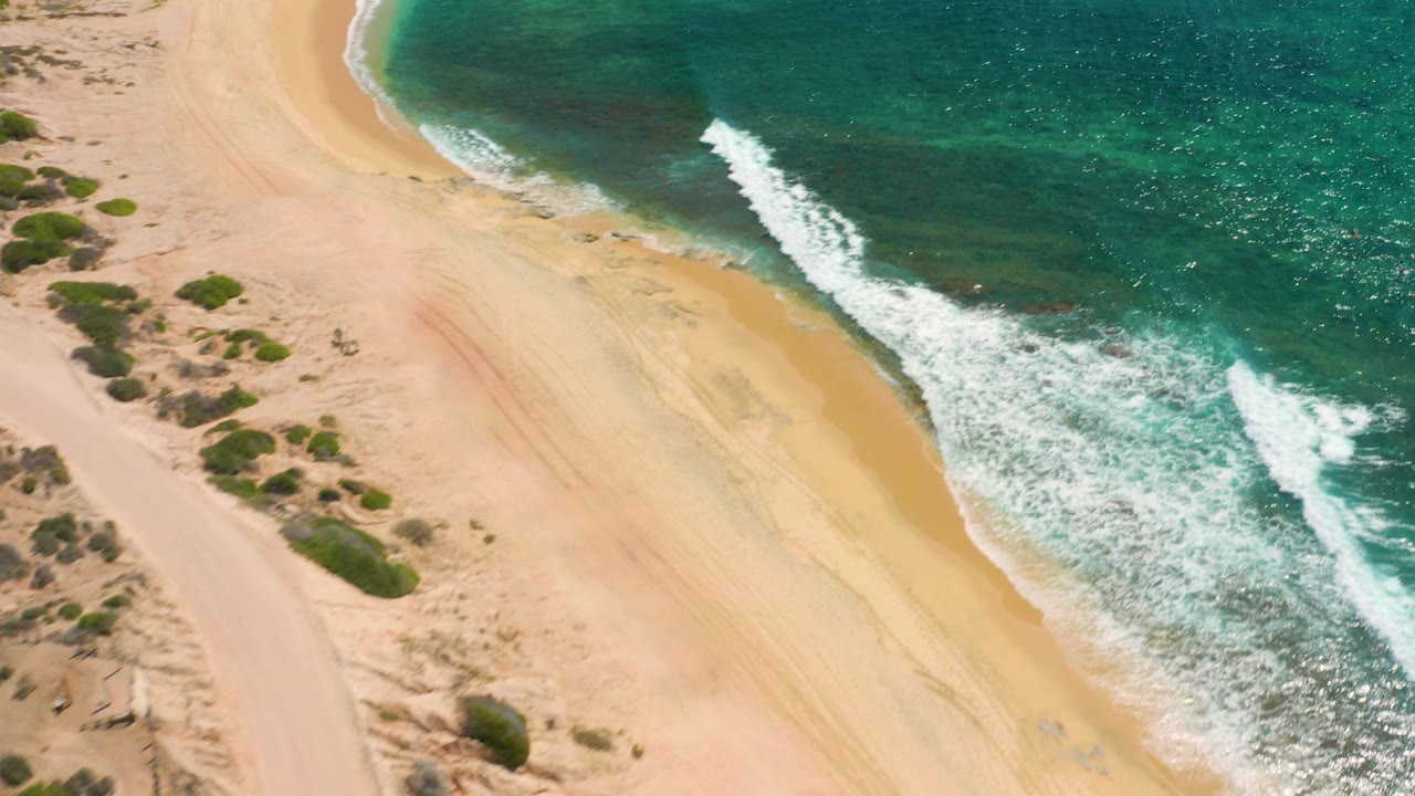 Aerial frame over Shipwrecks Beach in Baja California Sur with teal water and white surf curling onto empty tan sand, no visible people.