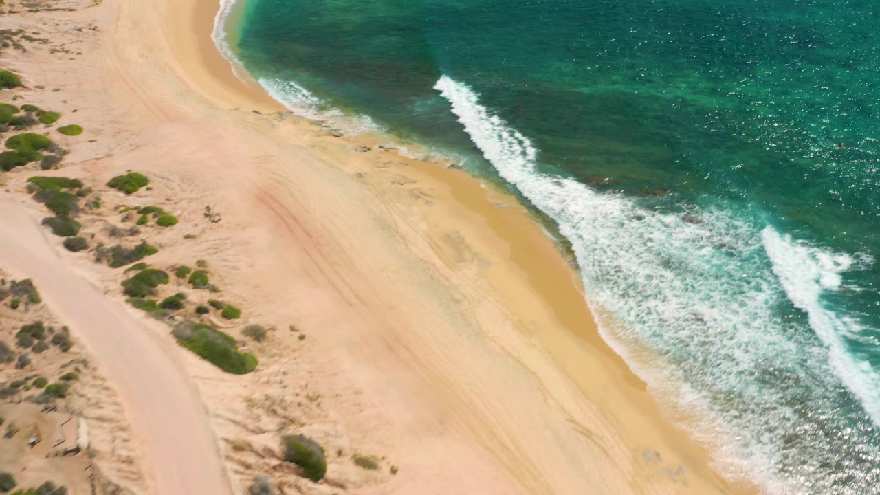 Overhead view at Shipwrecks Beach with a white wave line spreading across pale sand and green scrub set back from the water, no visible people.