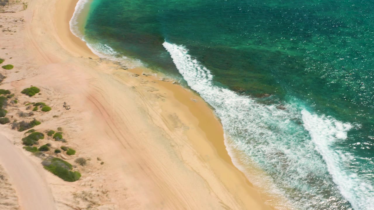 Aerial view of Shipwrecks Beach where the curved sand edge meets clear green-blue water and a narrow band of white surf, no visible people.