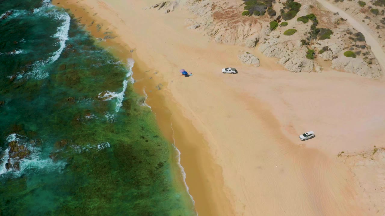 Aerial frame at Shipwrecks Beach, Baja California Sur, showing green-blue surf along pale sand, two parked light-colored cars, dunes at the top, and no visible people.