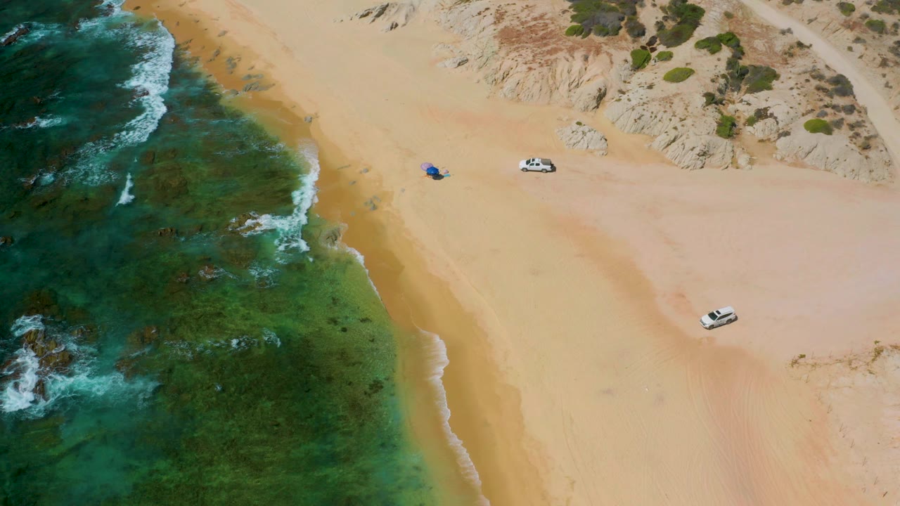 High aerial frame from Shipwrecks Beach, Baja California Sur, showing darker reef-colored water at left, broad open sand, two parked light vehicles, scrubby dunes, and no visible people.