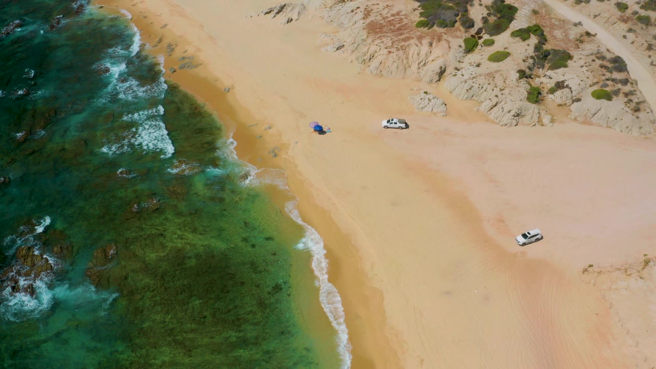 Aerial frame over Shipwrecks Beach in Baja California Sur showing green-blue surf at left, pale sand and dunes at right, two white vehicles on the beach, and no visible people.