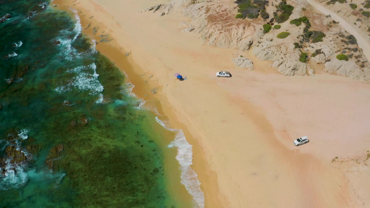 The drone angle over Shipwrecks Beach shows darker reef patches under the water, a broad strip of sand, two white vehicles, a blue spot on the beach, and no visible people.