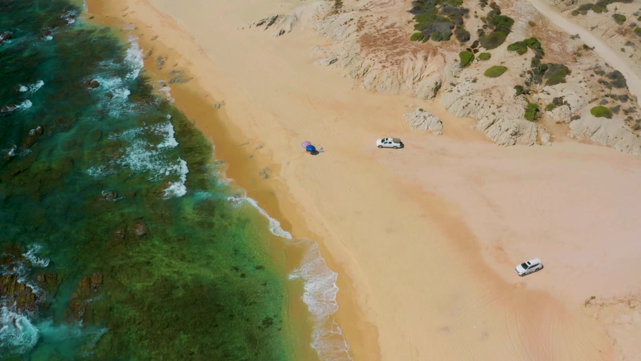 Mid-clip, the aerial view places foamy surf along the left edge, open sand across the center, two white vehicles near the dunes and lower beach, and no visible people.