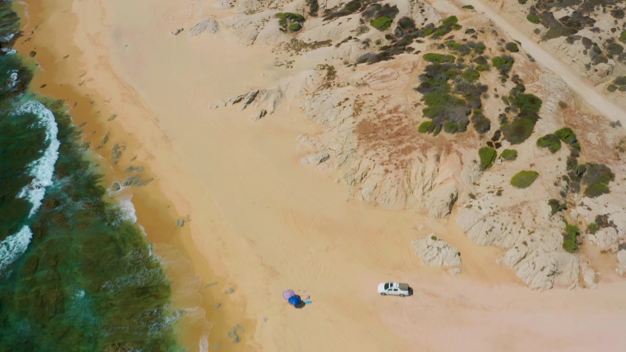 At 0:02, blue-green water and white wave foam border the left side of Shipwrecks Beach, with pale sand, tan rocks, scattered green shrubs, a white vehicle, and no visible people.