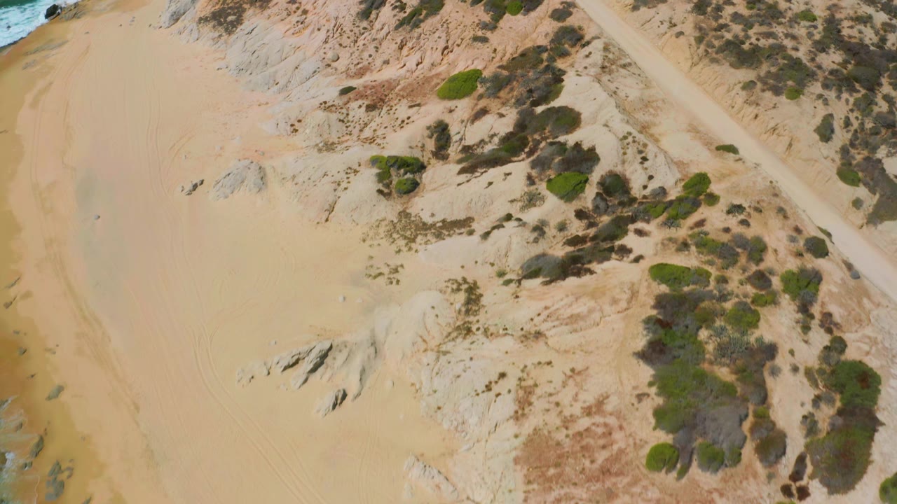 Bird's-eye frame over Shipwrecks Beach with dunes and scrub crossing the center, rock patches on the sand, a thin turquoise surf edge, and no visible people.
