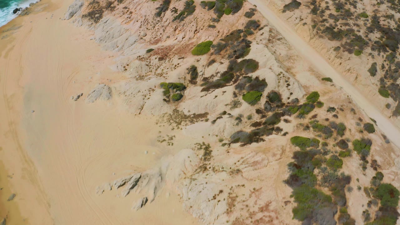 Top-down view of Shipwrecks Beach where a pale sandy track runs along scrub clusters and light rock outcrops, with no visible people in the frame.