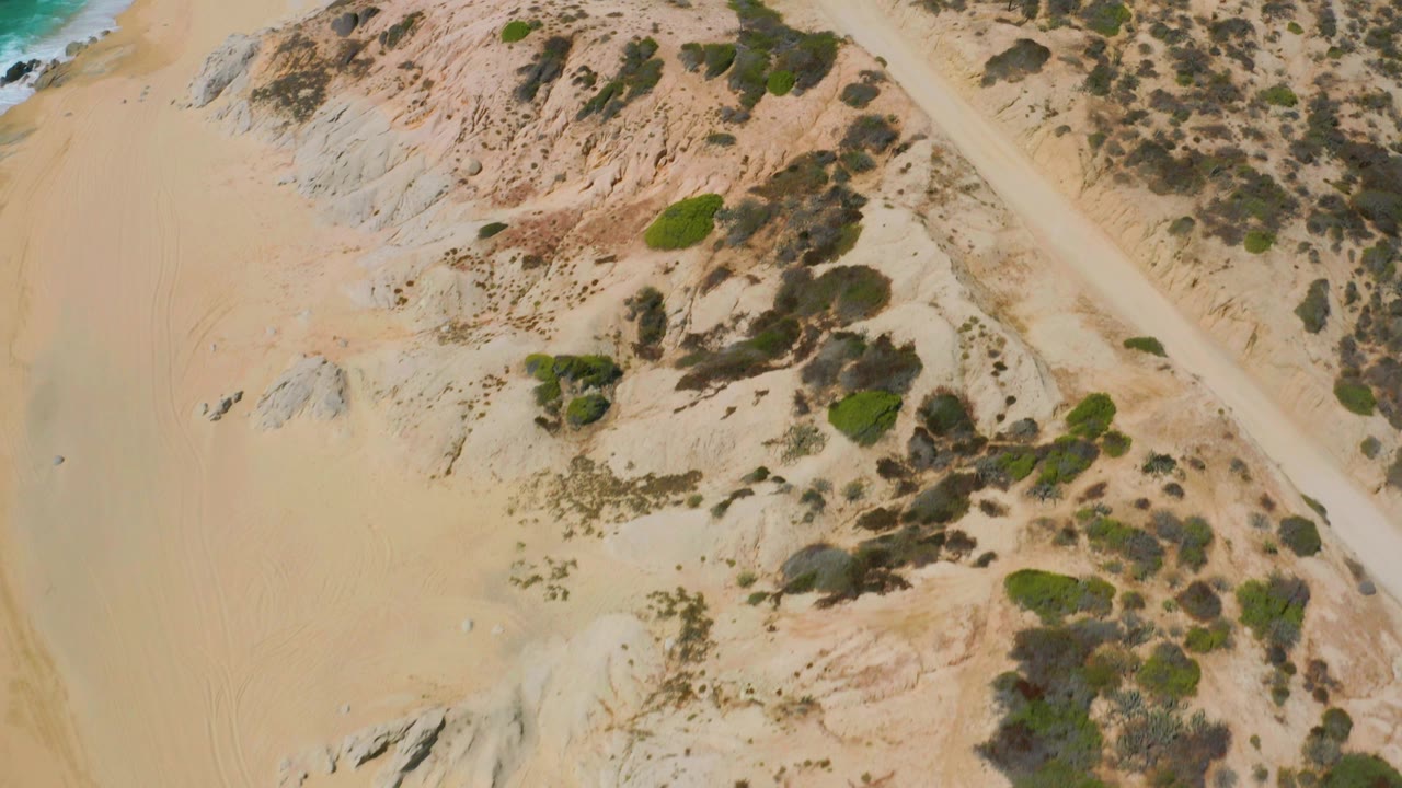 Aerial frame over Shipwrecks Beach showing open sand, clustered coastal scrub, pale rocks, faint curved tire marks, and no visible people.