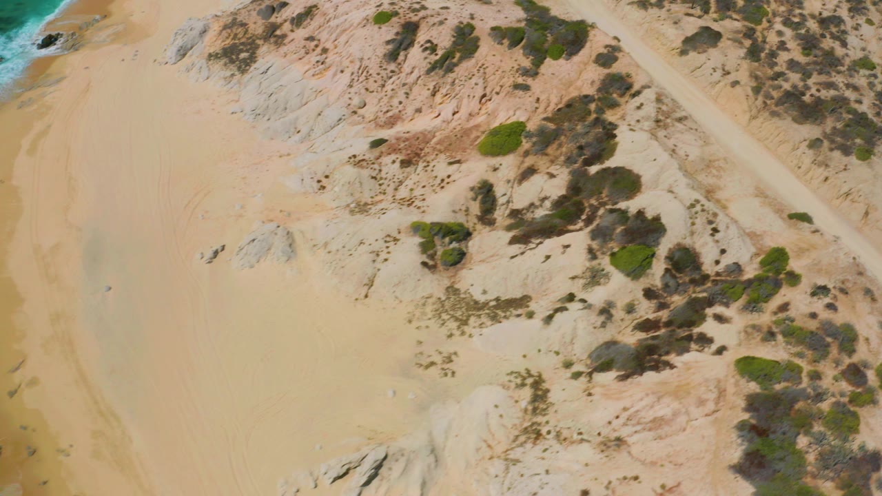 Overhead view of Shipwrecks Beach with pale sand, low dunes, scattered rocks, scrub vegetation, and a strip of turquoise surf at the upper left, with no visible people.