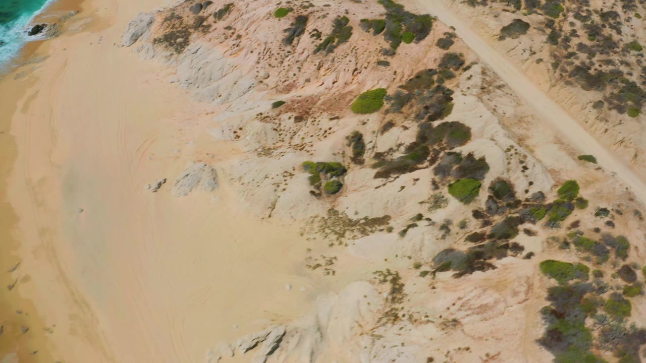 Aerial frame at Shipwrecks Beach showing broad sand, clustered green scrub, exposed rock patches, and white foam along the right shoreline, with no visible people.