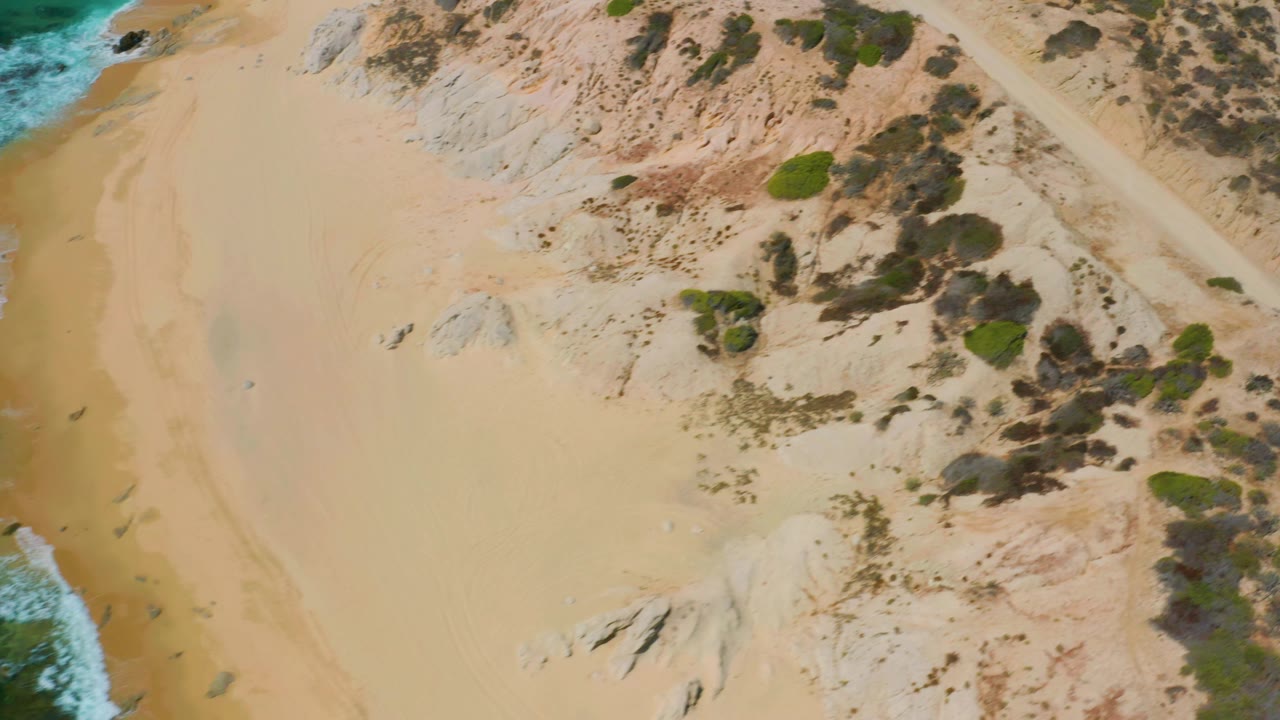 Top-down view of Shipwrecks Beach with a large open sand area, low scrub, small rock outcrops, and surf edging into the frame, with no visible people.