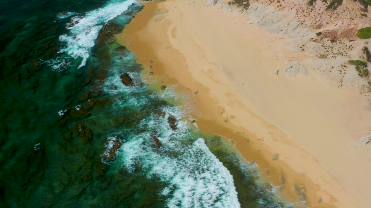 Aerial view at Shipwrecks Beach, Baja California Sur, where a thicker band of white foam curls along the beach over submerged rocks, no visible people.