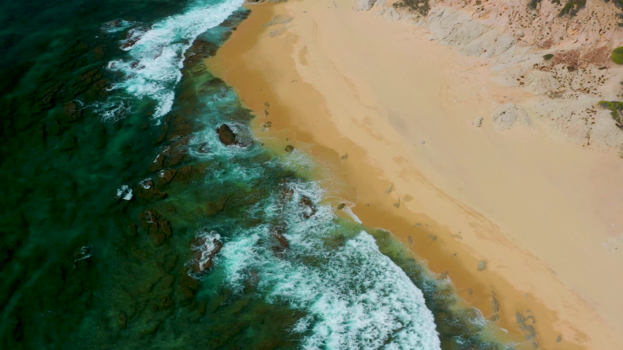 Drone-style overhead frame from Shipwrecks Beach, Baja California Sur, with dark green water on the left, a white surf line, and broad golden sand on the right, no visible people.