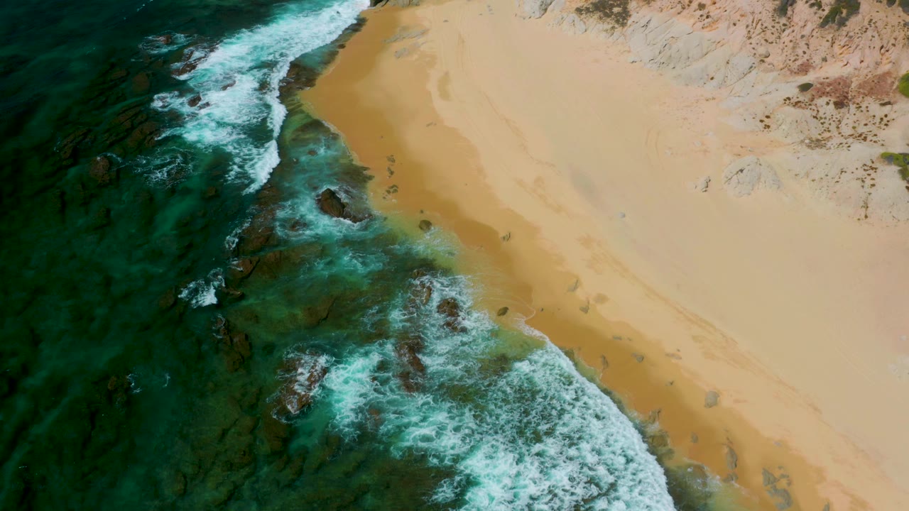 Aerial frame at Shipwrecks Beach, Baja California Sur, showing foam spread farther across the shallow rocky water while the golden beach stays empty, no visible people.