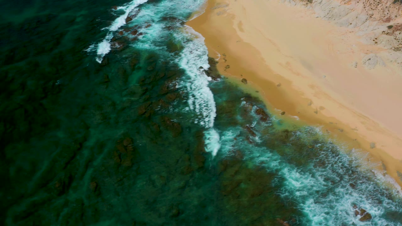 Aerial view at Shipwrecks Beach, Baja California Sur, where white surf curls over submerged rocks below a tan sandy beach, with no visible people.