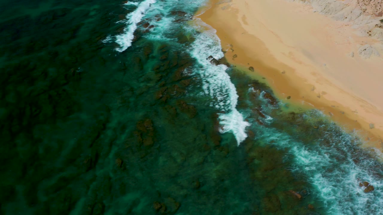 Top-down aerial frame at Shipwrecks Beach, Baja California Sur, showing a bright wave line running diagonally through green-blue water toward the sandy edge, with no visible people.