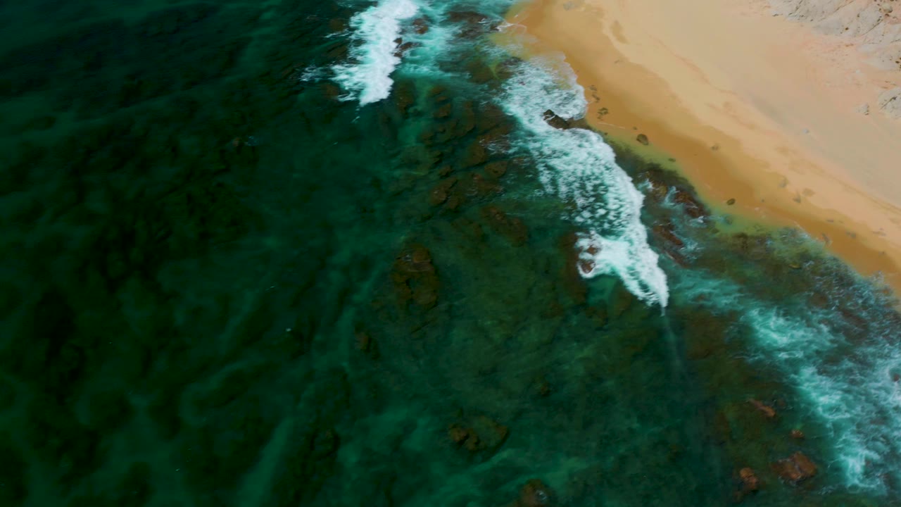 Aerial frame at Shipwrecks Beach, Baja California Sur, with deeper green water filling the left side, surf breaking across center, sand at the upper right, and no visible people.