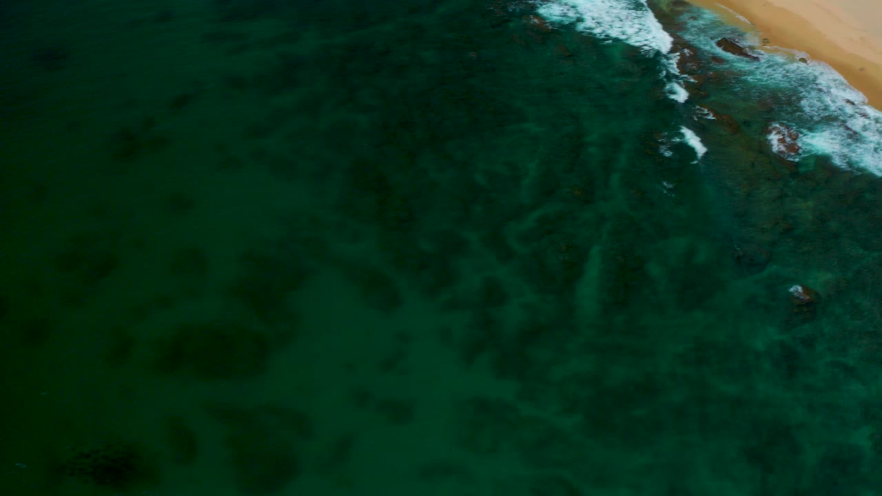 High aerial frame over Shipwrecks Beach, Baja California Sur, with dark green water, pale submerged sand patterns, white surf at the top edge, and no visible people.