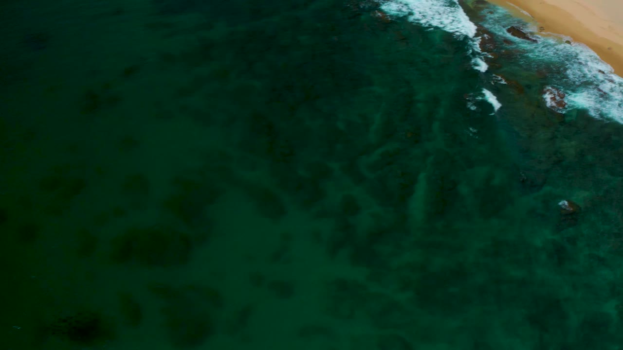 Drone-like overhead view at Shipwrecks Beach showing green-blue water, broken white wave foam near the upper beach edge, faint underwater ripples, and no visible people.