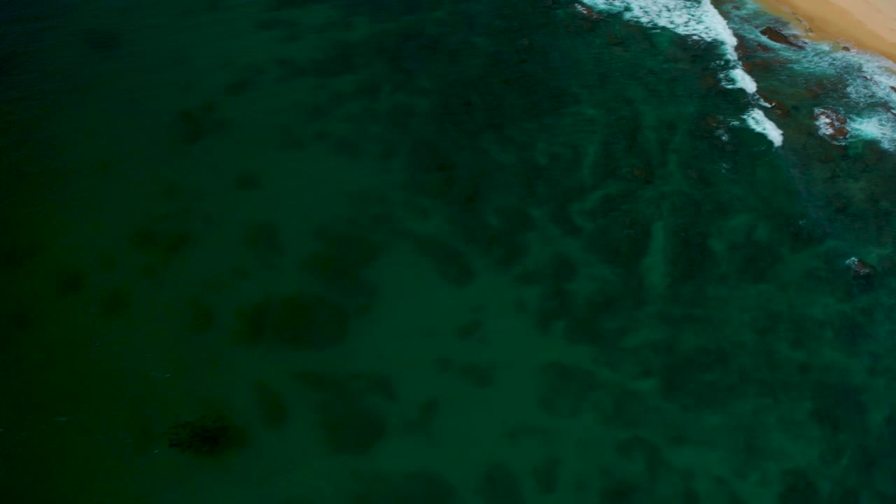 Aerial frame from Shipwrecks Beach with the sandy beach mostly cropped to the upper right, white surf crossing the top edge, dark underwater patches, and no visible people.