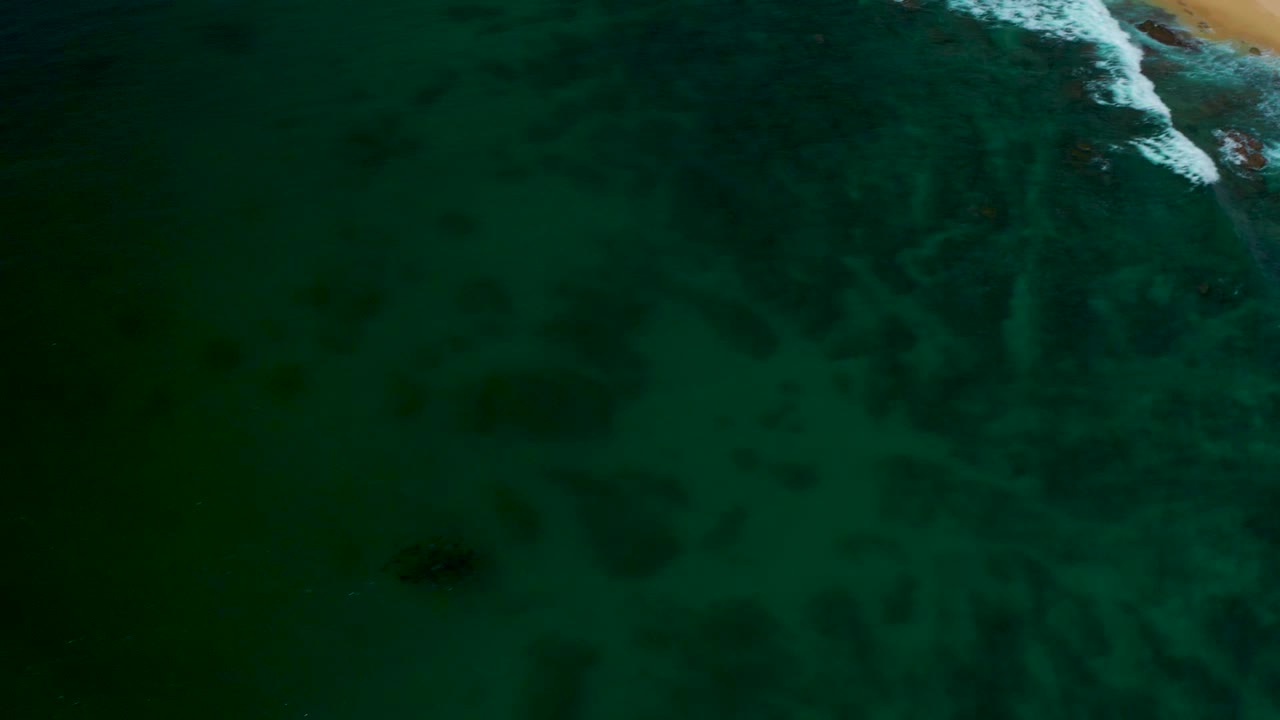 Overhead view at Shipwrecks Beach showing dark green water, faint pale sand trails under the surface, a small white wave break at the upper right, and no visible people.