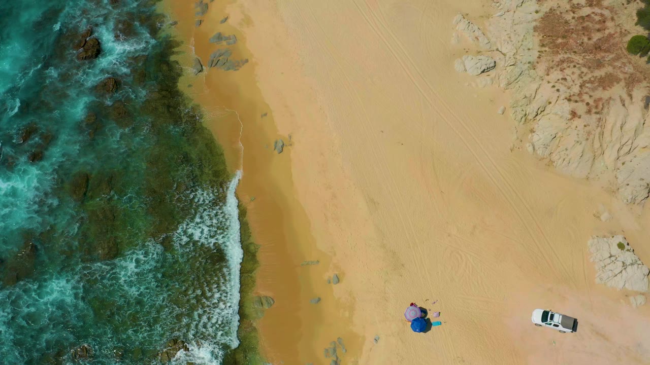 Top-down aerial frame at Shipwrecks Beach, Mexico, showing blue-green water over reef on the left, wide tan sand, pale rock at upper right, a white vehicle, small colorful beach items, and no visible people.