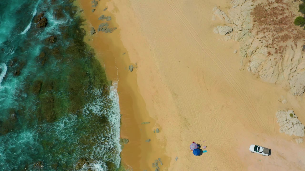 At 0:02, the overhead Shipwrecks Beach view shows more foamy water along the reef edge, broad tan sand, the white vehicle near the lower right, small beach items near the lower center, and no visible people.