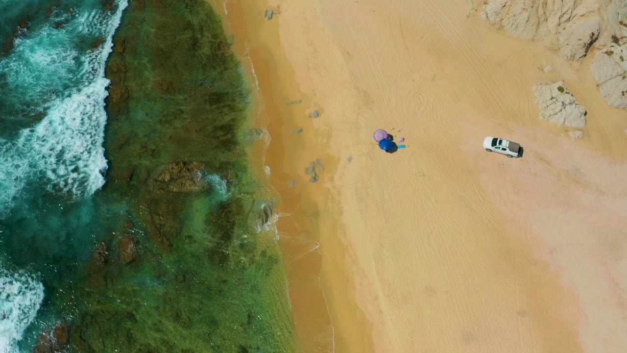 Overhead view at Shipwrecks Beach, Baja California Sur with teal surf on the left, tan sand on the right, a white vehicle, and distant people beside a blue umbrella.