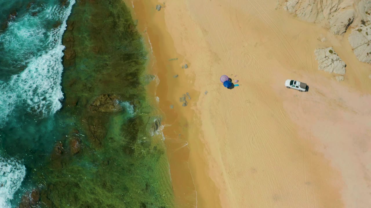 Aerial frame over Shipwrecks Beach showing breaking white surf, green shallows, open sand, a white vehicle, and distant people gathered near a blue umbrella.