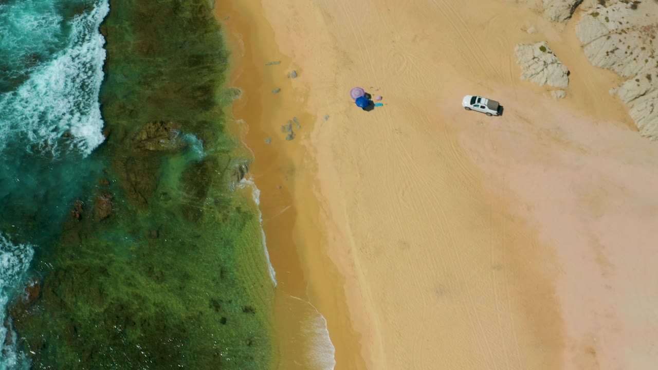 Top-down view at Shipwrecks Beach with surf and dark reef patches along the left, open sand across the right, a white vehicle, and distant people near a blue umbrella.