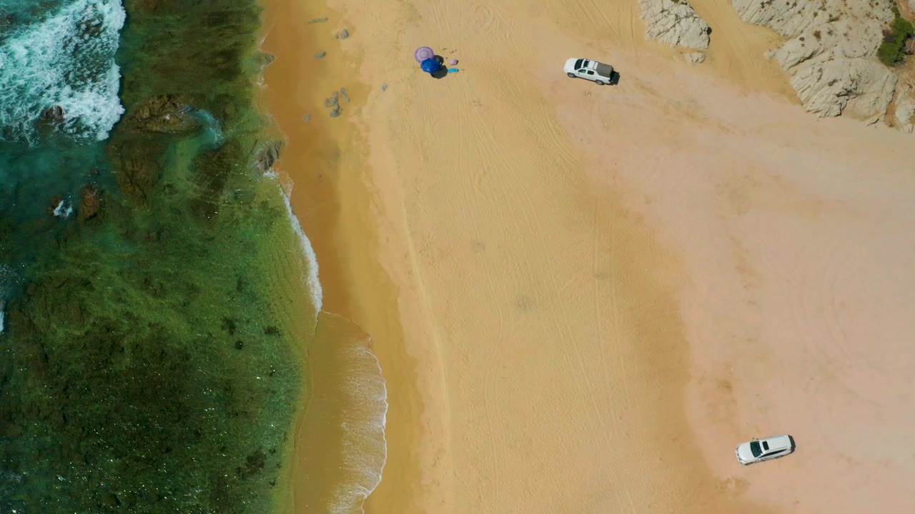 Aerial shoreline view with foamy surf, sand, rocks, a white vehicle, and distant umbrella.