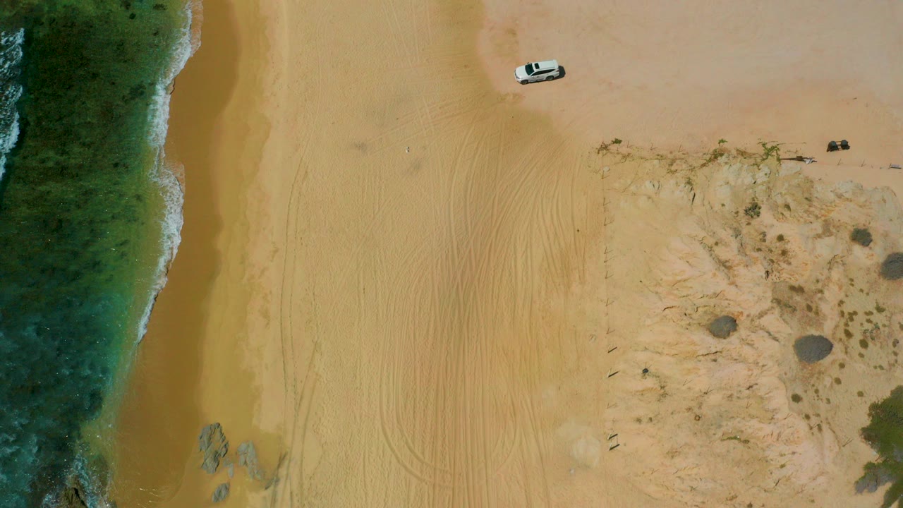 Overhead beach with surf, sand, rocks, and a small white vehicle.