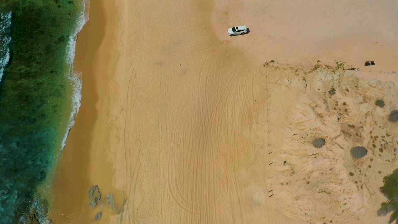 Aerial frame over Shipwrecks Beach with the ocean edge running down the left, broad tan sand marked by faint tire tracks, scrubby beach vegetation on the right, a small white vehicle near the top, and no visible people.