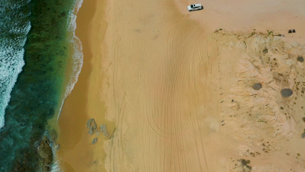 Top-down view of Shipwrecks Beach showing a small white vehicle near the upper sand, curving tire marks across the beach, darker rocky water at left, low scrub at right, and no visible people.