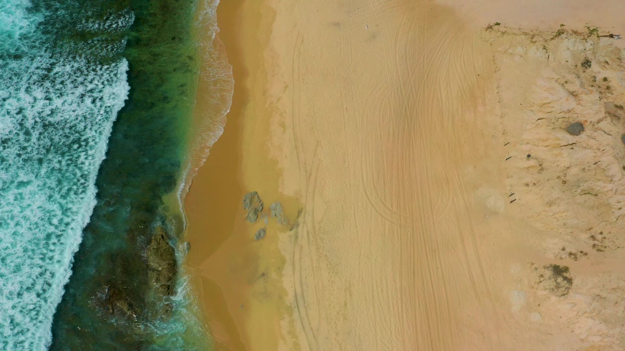 Overhead frame at Shipwrecks Beach with foamy surf pushing in from the left, a narrow wet-sand strip, pale dry sand with track marks, scattered shoreline rocks, scrub to the right, and no visible people.