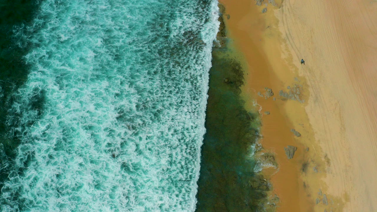 Overhead view of green-white surf pressing against a narrow dark shoreline band and tan sand at Shipwrecks Beach, Baja California Sur, with no clearly visible people.