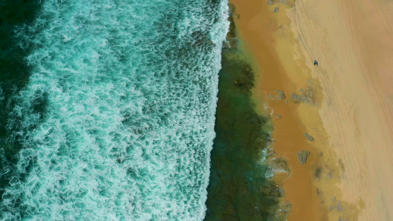 A dense field of white foam covers the left half of the frame while tan beach and scattered rocks remain visible on the right at Shipwrecks Beach, Baja California Sur.