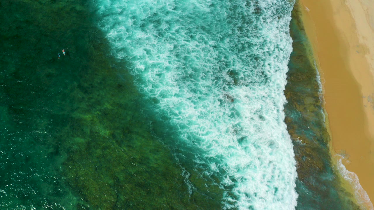 Overhead view at Shipwrecks Beach, Baja California Sur, with green-blue water, white surf, a strip of sand on the right, dark underwater patches, and no visible people.