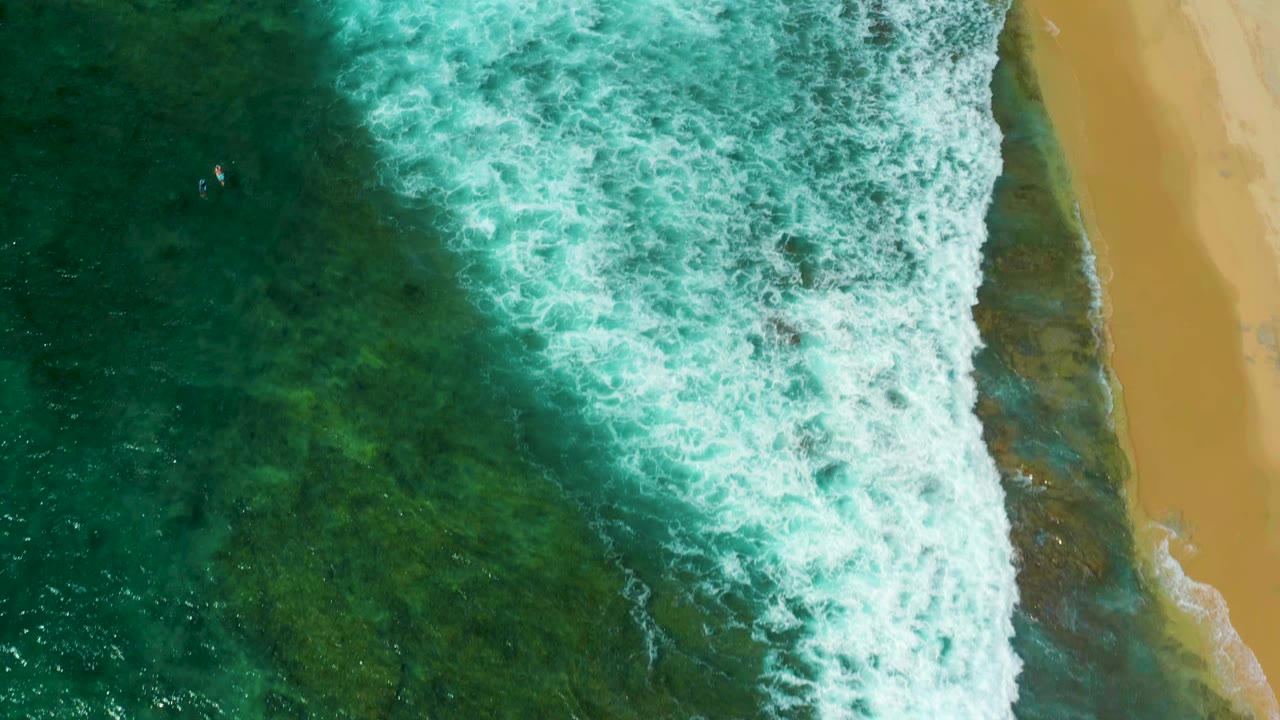 Aerial frame from Shipwrecks Beach showing a thick band of white foam over green-blue shallows with golden sand along the right edge and no visible people.